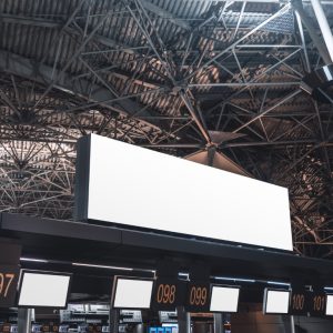 An empty white advertising or information billboard mock-up on the top of check-in and baggage claim area of a modern airport terminal, with multiple numbered counters and blank info screens below