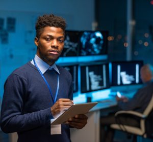 Young serious African programmer with tablet standing in front of camera against colleague by workplace